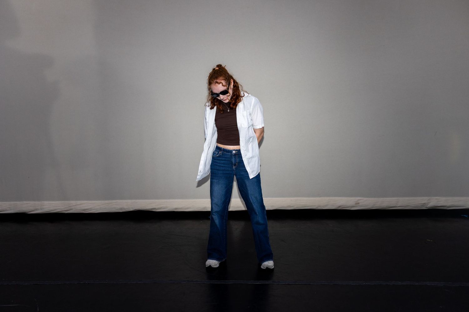 Woman in white coat and flared jeans laughing expressively on a dark performance stage floor.