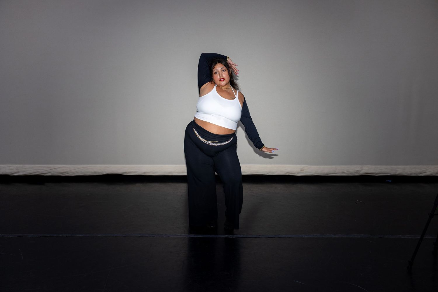 A woman in a white crop top and navy pants strikes an expressive dance pose against a gray studio backdrop.