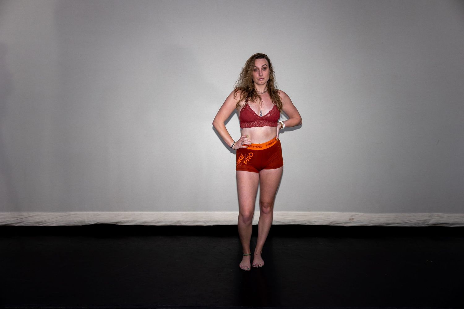 Woman in red sports bra and shorts stands confidently against gray wall on black floor.
