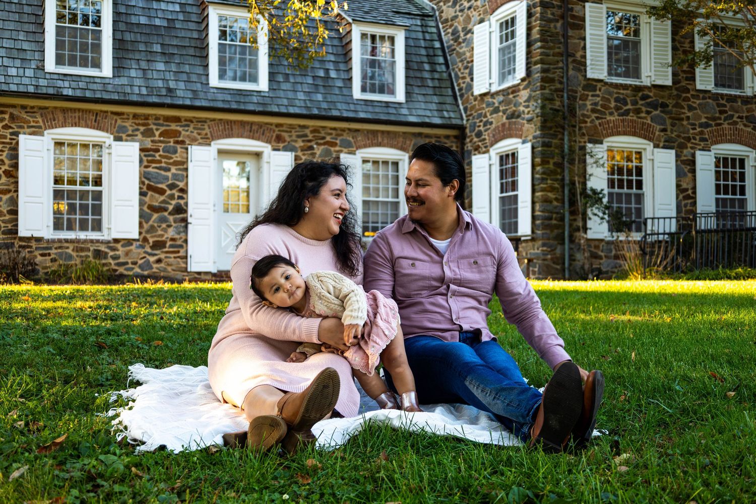 Happy family of three sitting on grass in front of a stone house, sharing a joyful moment outdoors.