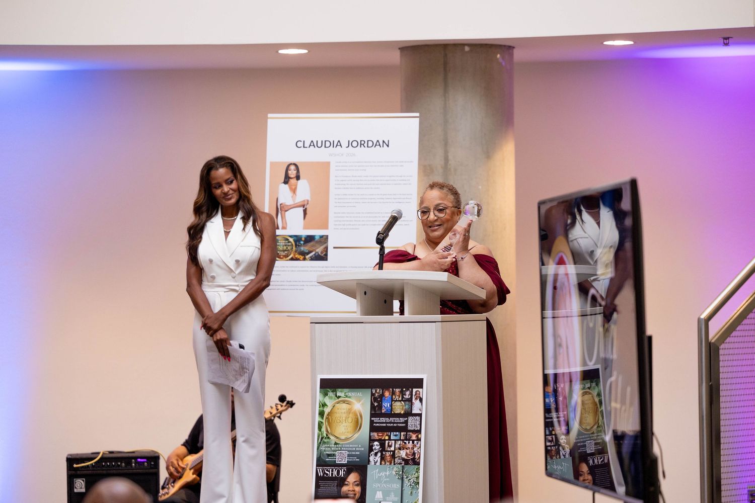 Claudia Jordan honored at an awards ceremony, standing at a podium with a trophy in an elegant venue.