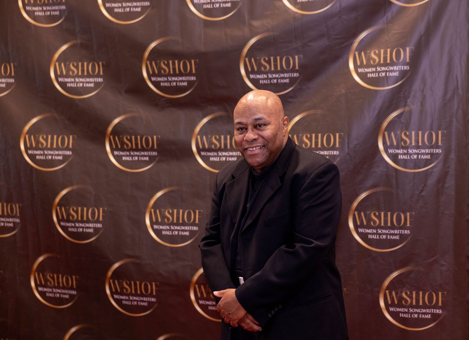 Man smiling in black outfit at WSHOF Women Songwriters Hall of Fame event backdrop.