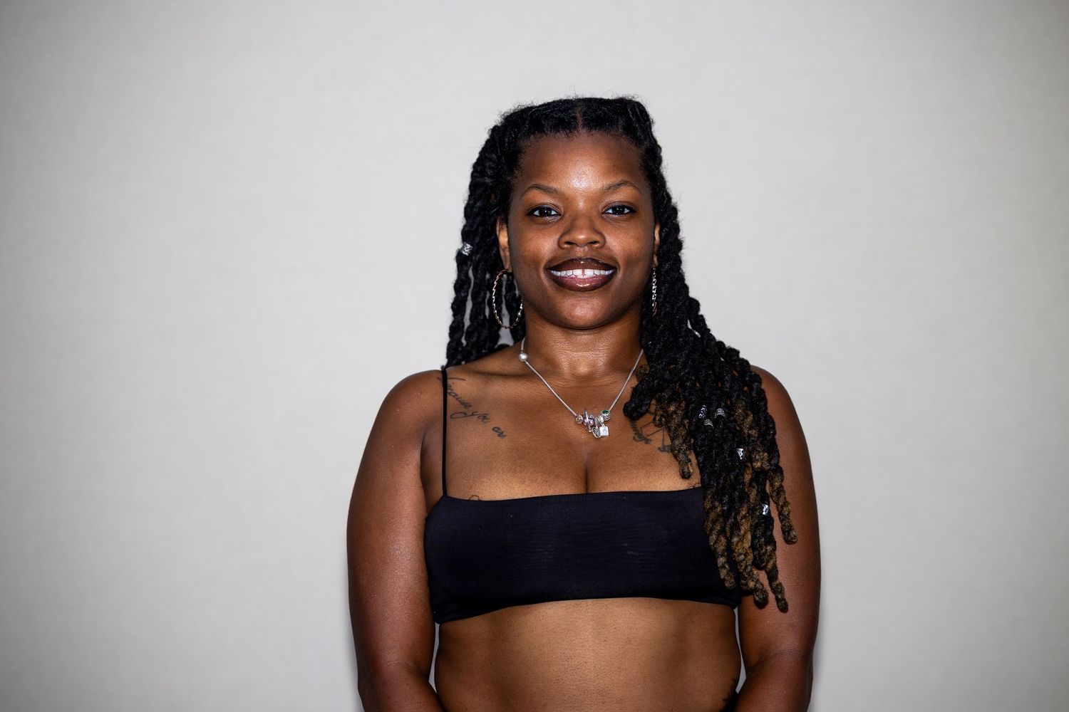 A woman with braided locs smiling warmly, wearing a black bandeau top and silver necklace against a white background.