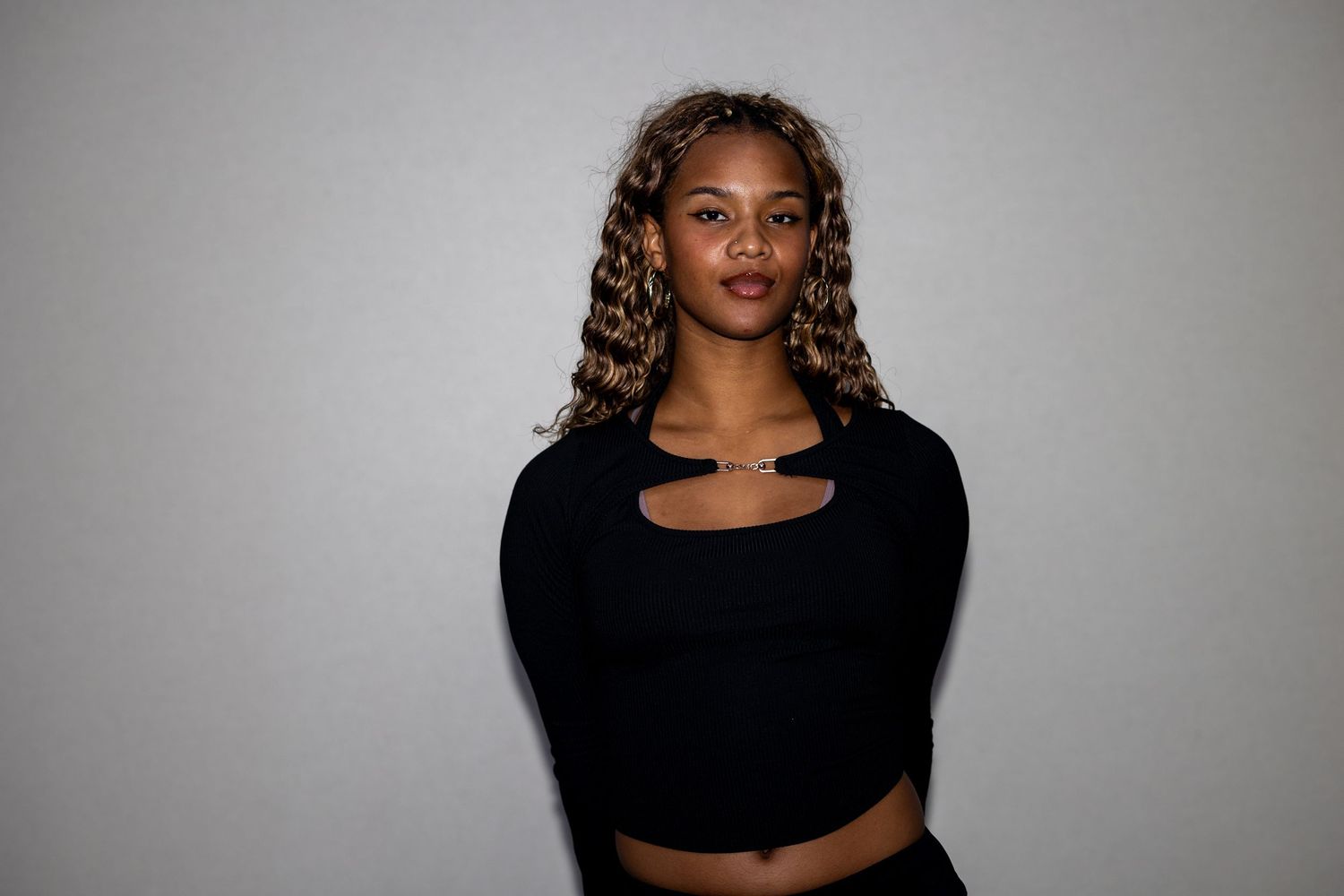 Young woman in black cutout crop top with highlighted curly hair poses against neutral background.