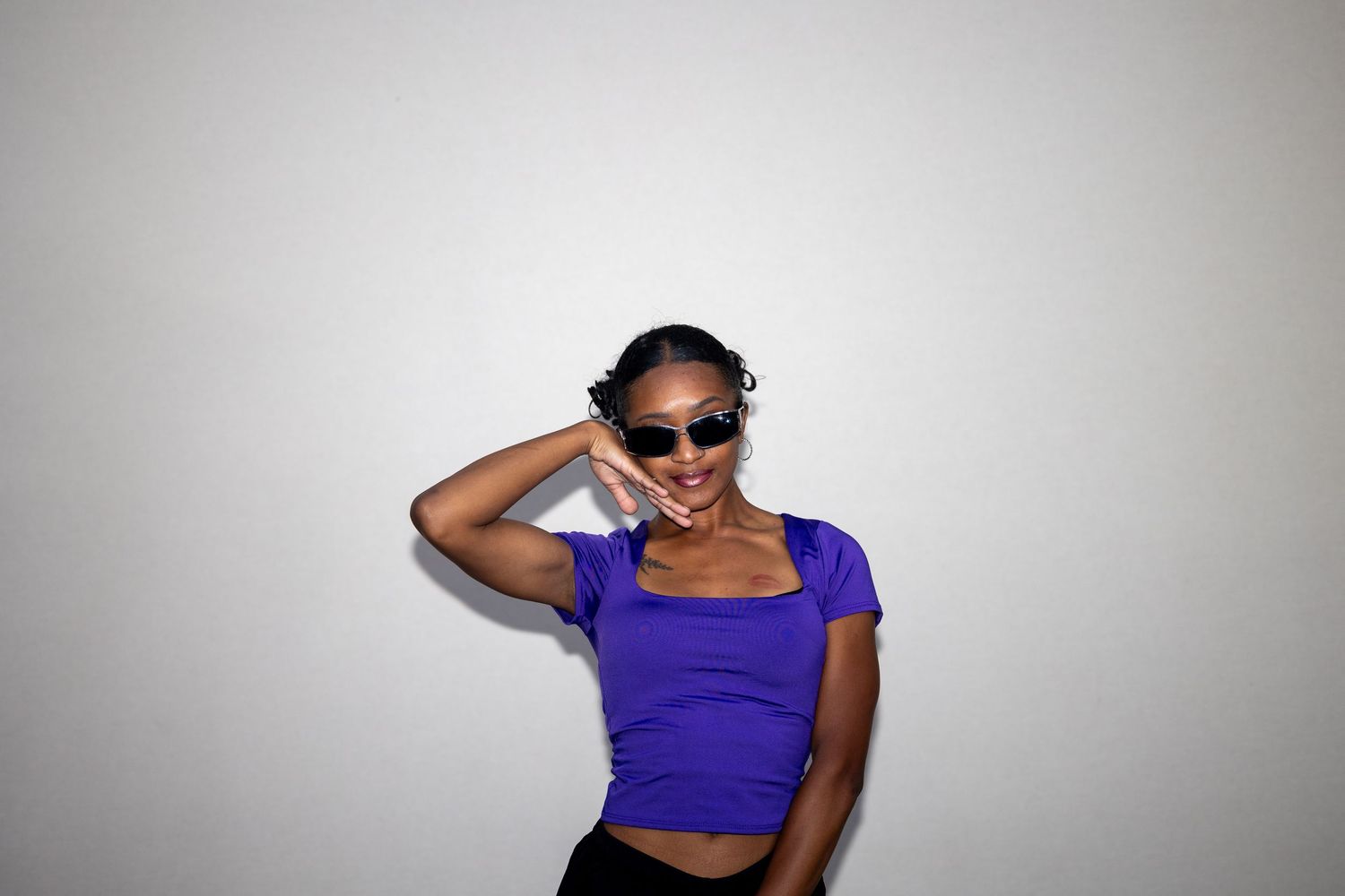 Young Black woman in a purple crop top and sunglasses posing with hand raised near her head indoors.