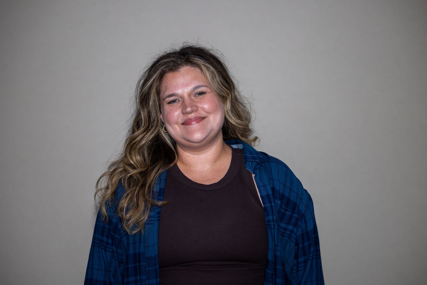 Smiling young woman with wavy blonde hair wearing a blue flannel shirt over a dark top against a gray background.