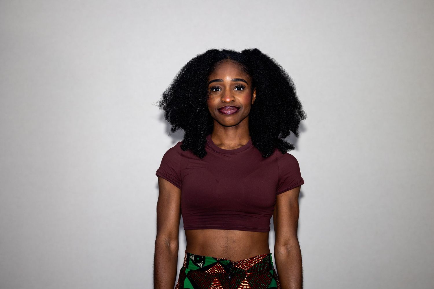 Young woman in maroon crop top and colorful skirt smiling against white background.