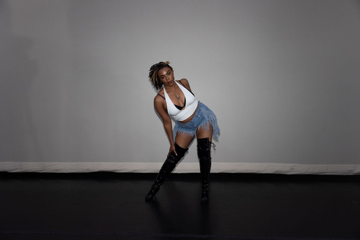 Woman in white crop top and denim mini skirt leans forward in thigh-high boots in studio.