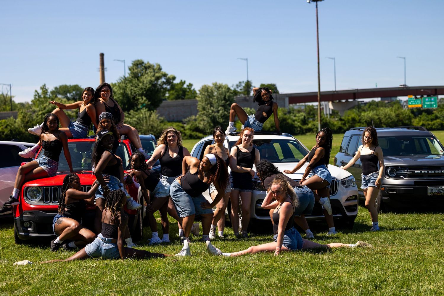 Group of young women posing together outdoors near parked cars on a sunny day.