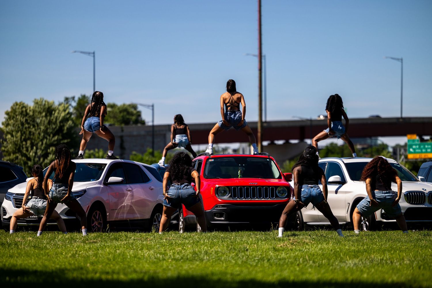 Women dancing on and around cars outdoors on a sunny day in an open grassy area.