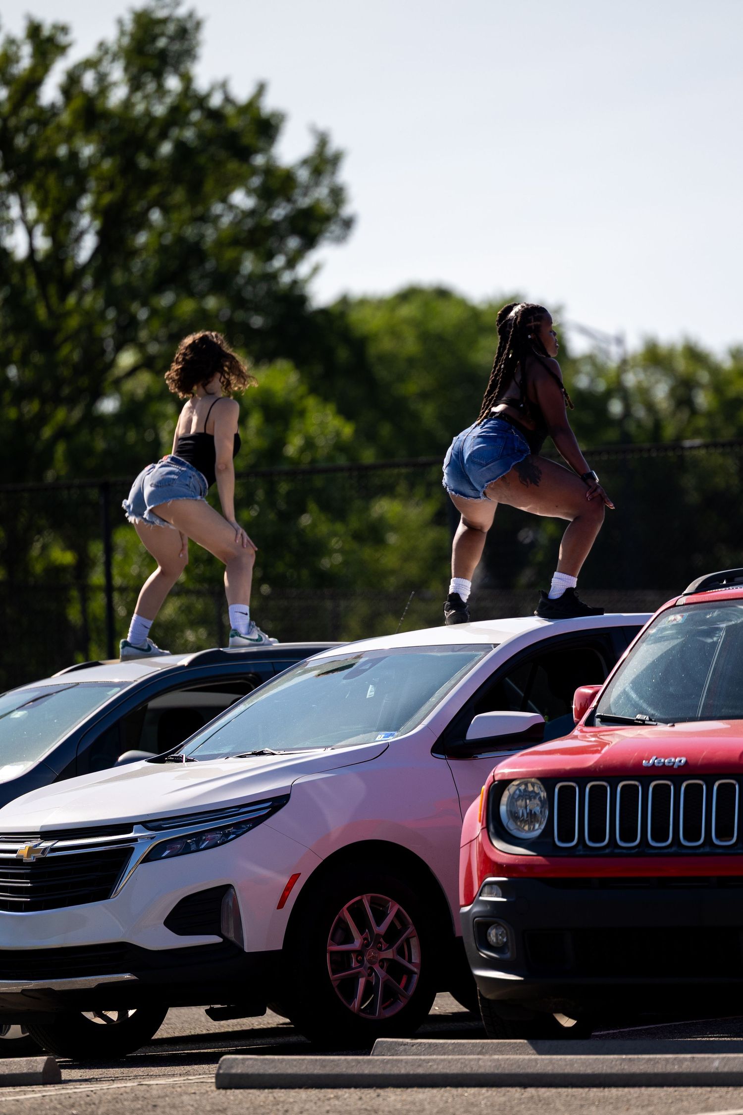 Two women in denim shorts dancing on top of cars in a parking lot on a sunny day.
