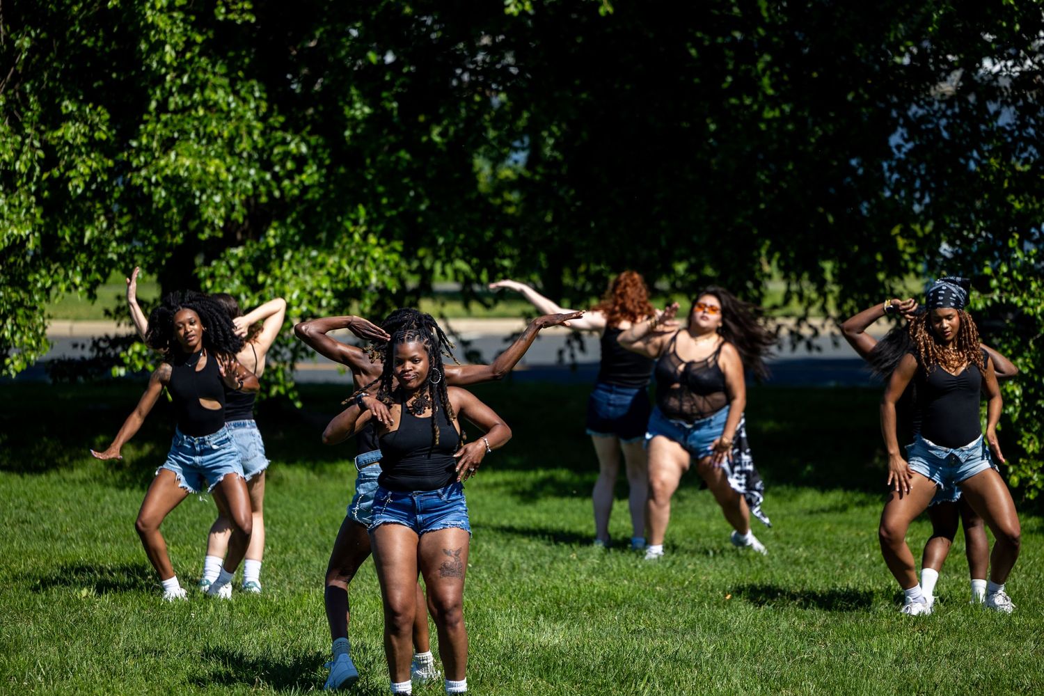 A group of young women dancing energetically on green grass in a sunny park surrounded by trees.