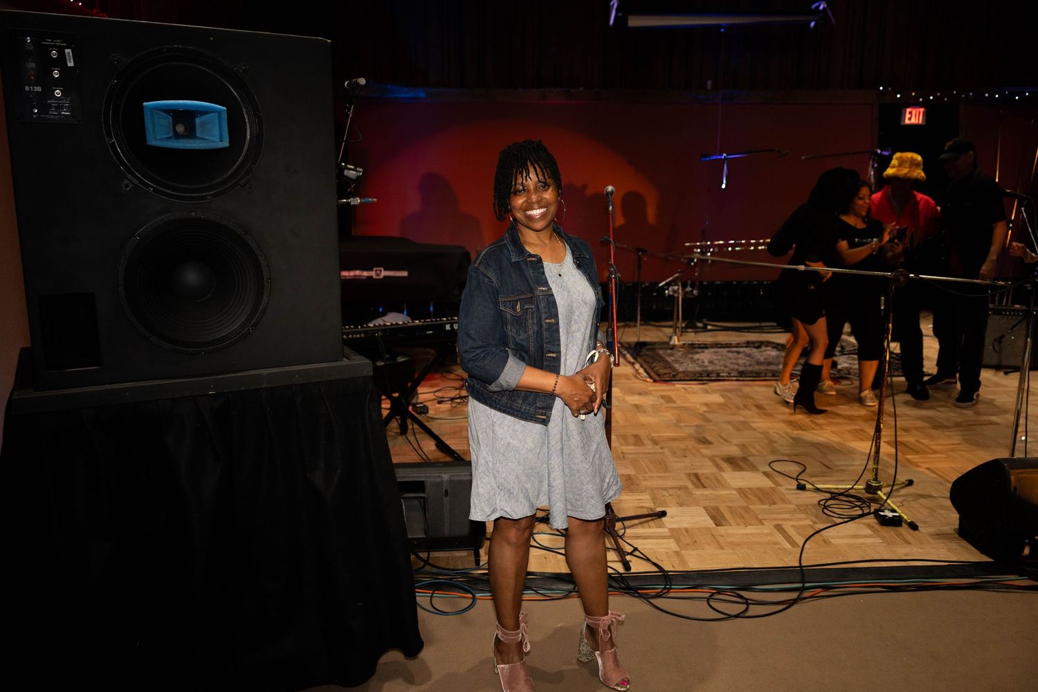 A smiling woman in a denim jacket and white dress stands next to a large speaker in a music venue.