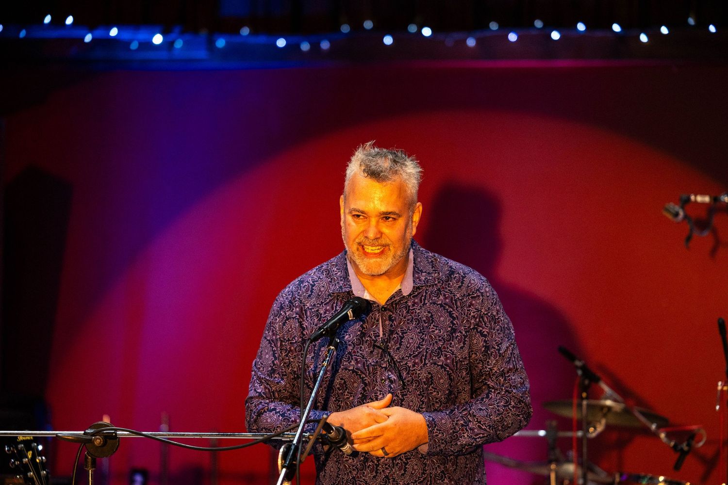 Man speaking at microphone on stage with colorful red and blue lighting at an indoor event.