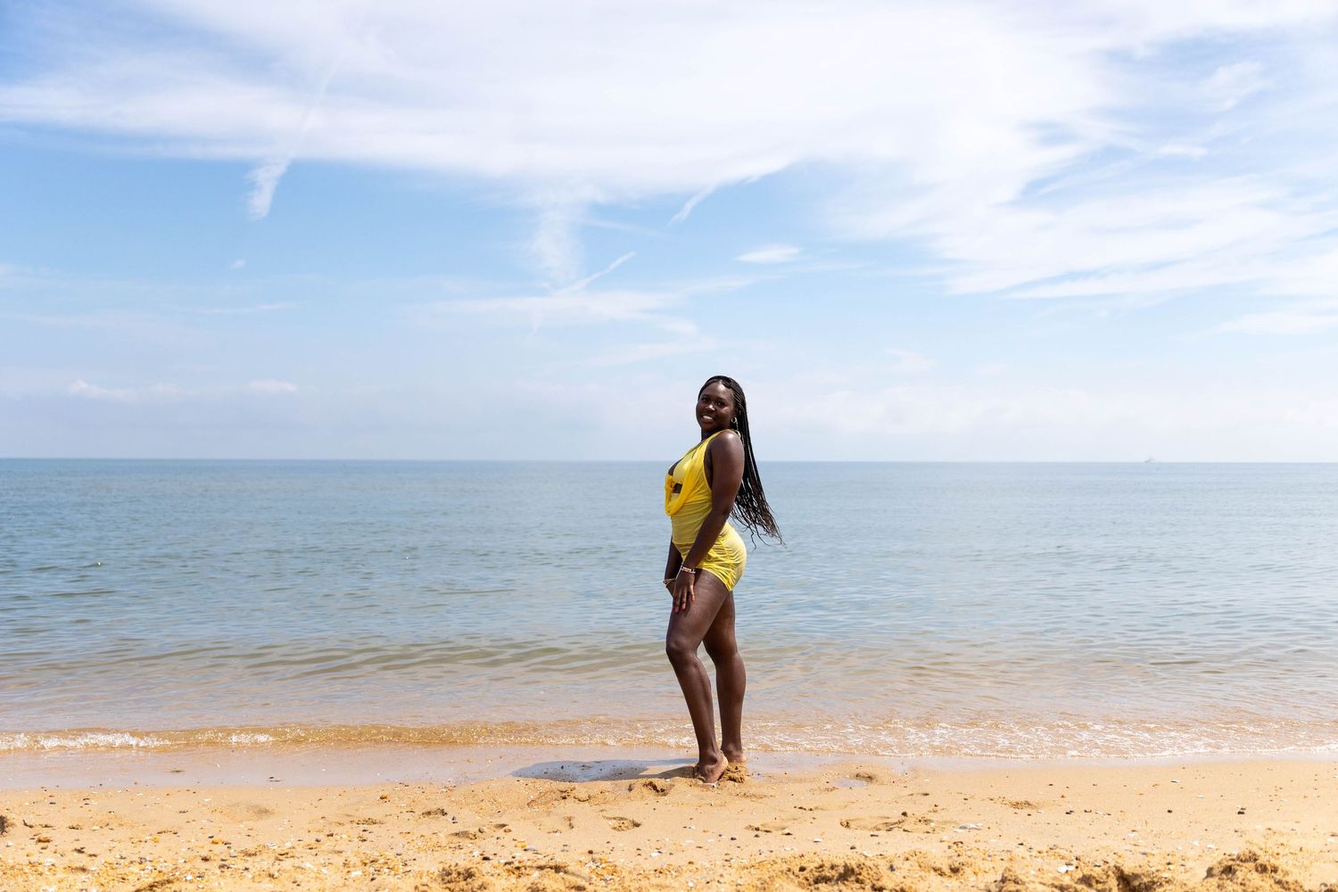 Woman in yellow swimsuit posing on sandy beach with calm blue ocean and sky backdrop.
