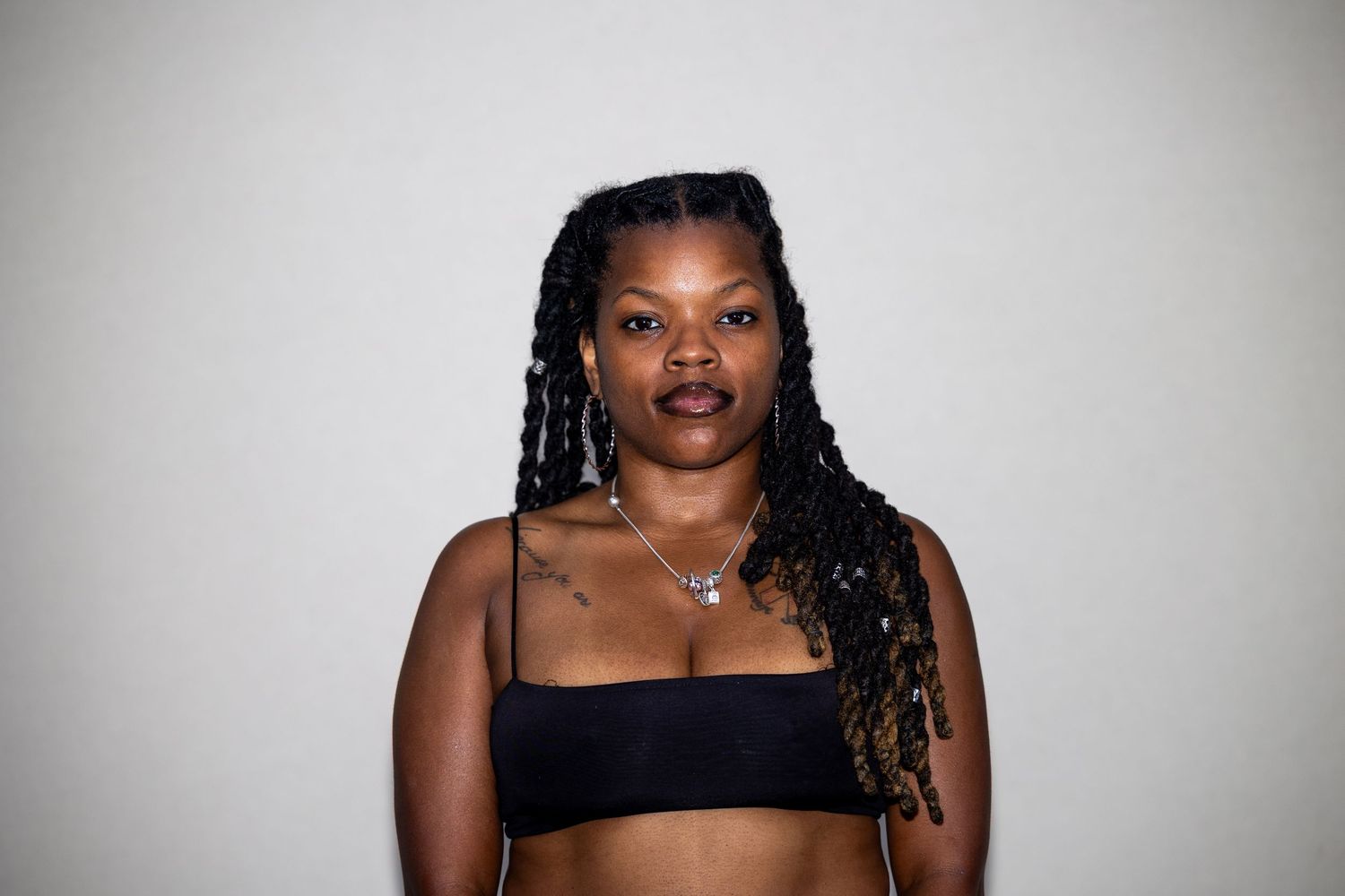 Woman with long braids wearing a black crop top and silver jewelry, posing against a white background.
