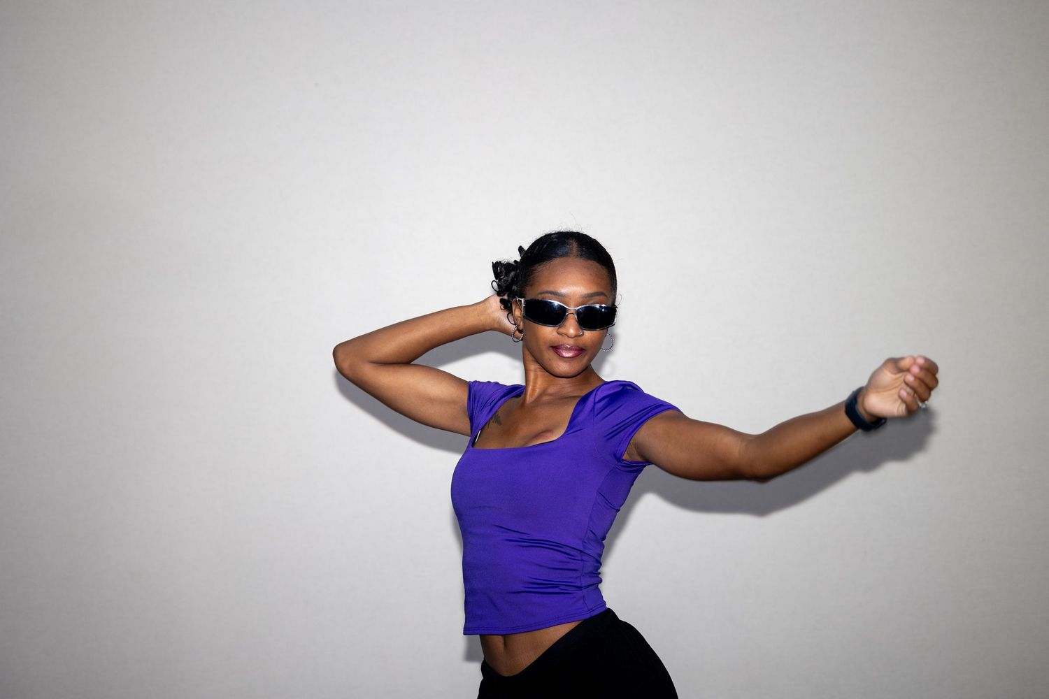Black woman in purple crop top and sunglasses dances energetically against a gray background.