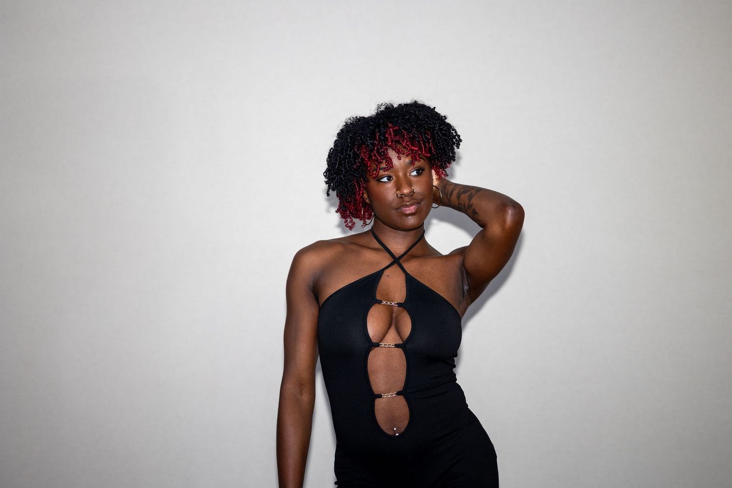 Woman with red-tipped curly hair in black cutout halter dress posing against light gray background.