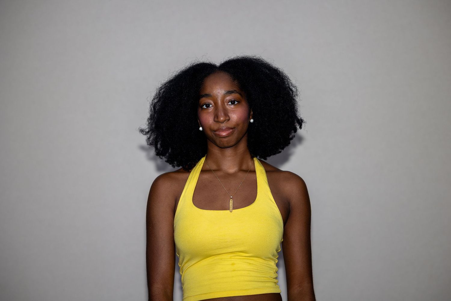 A slim woman with a natural afro wears a yellow halter crop top and pearl earrings against a gray background.