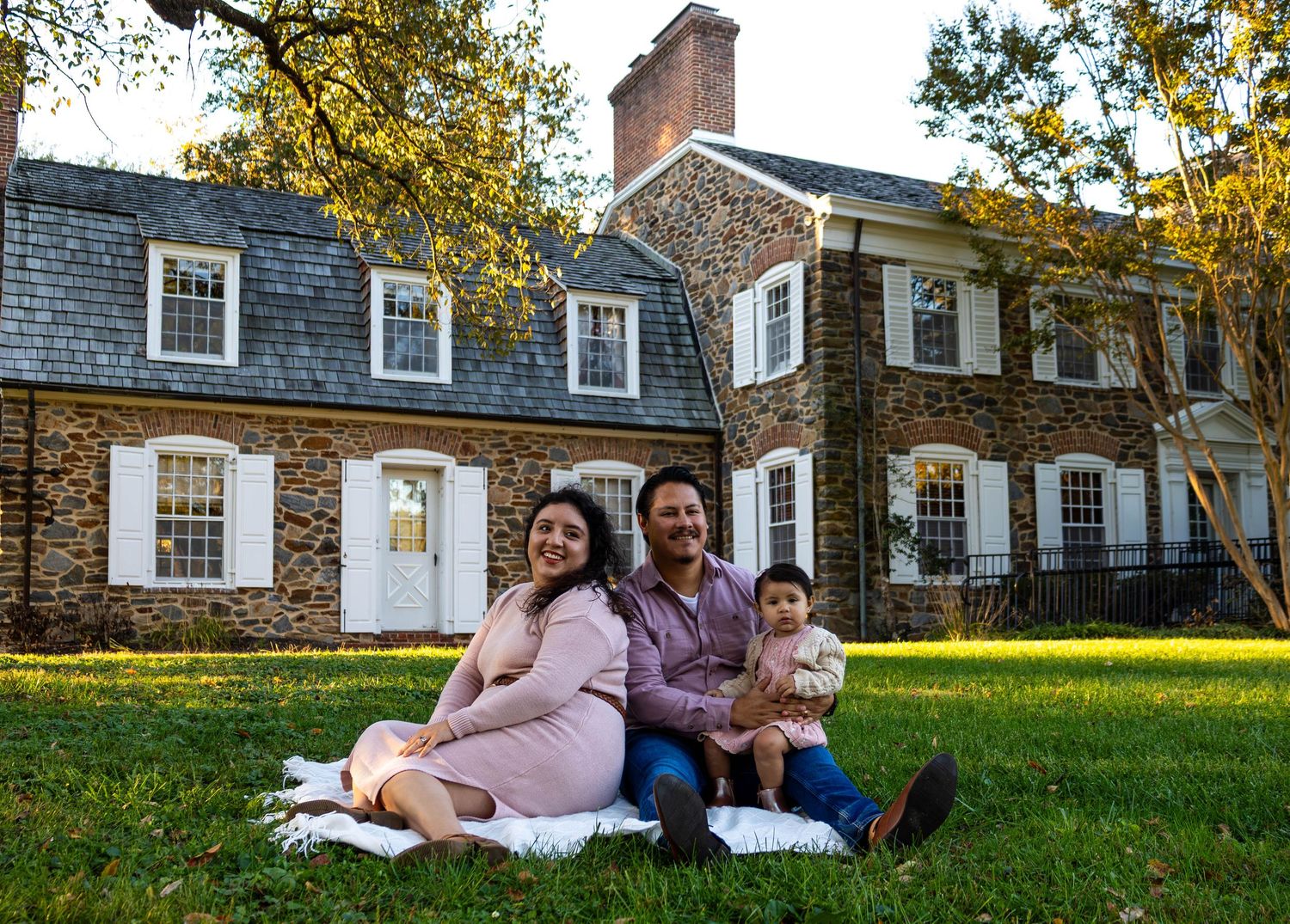 Family of three sitting on grass in front of a historic stone house, smiling for a portrait photo.