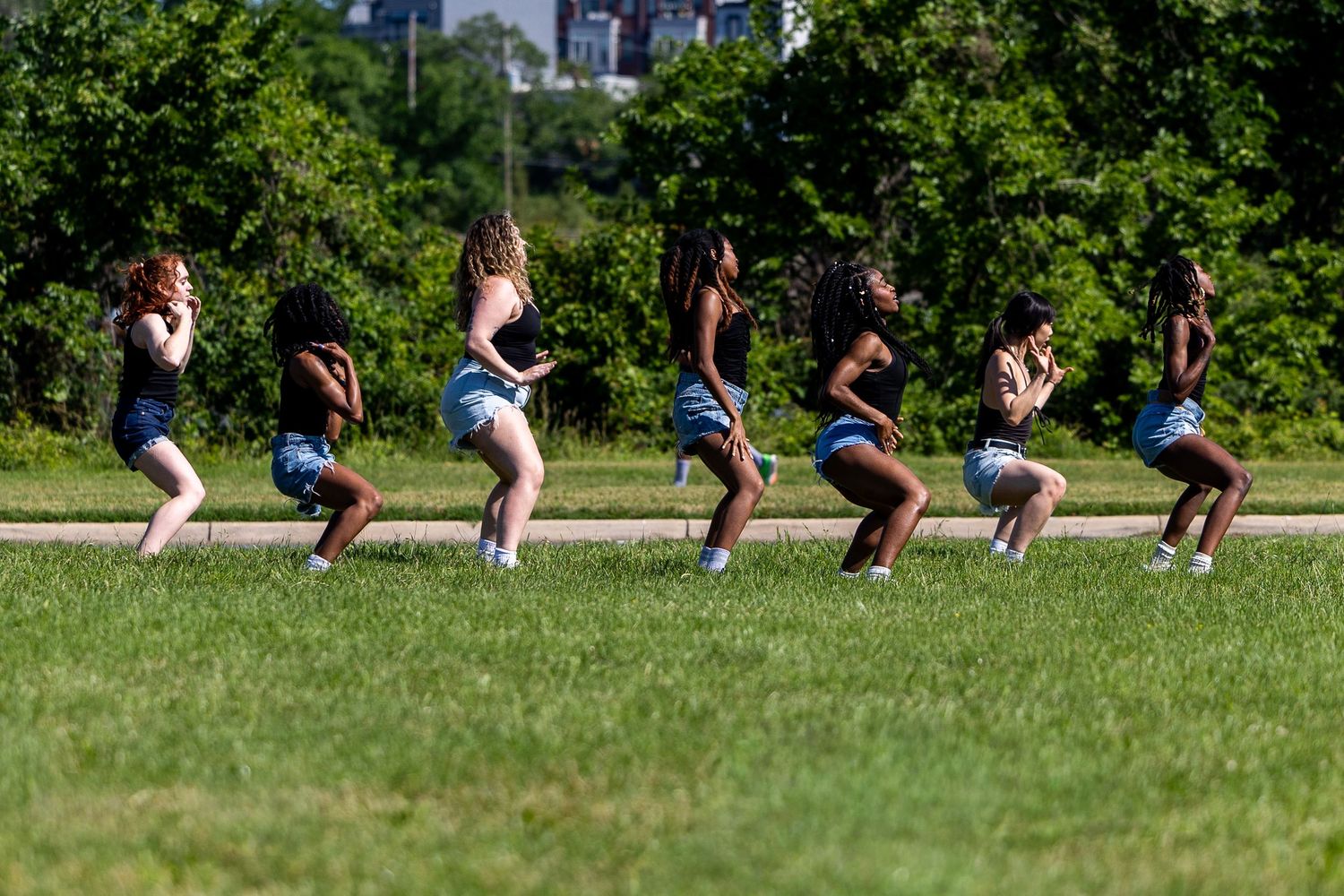 Six young women dancing in sync on a green grass field outdoors on a sunny day.