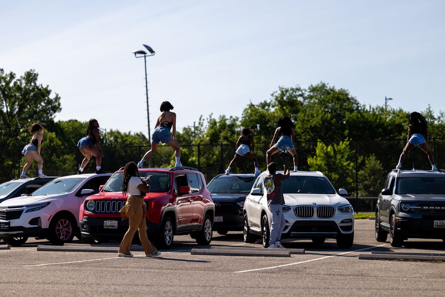 People standing on top of cars in a sunny parking lot surrounded by trees and various vehicles.