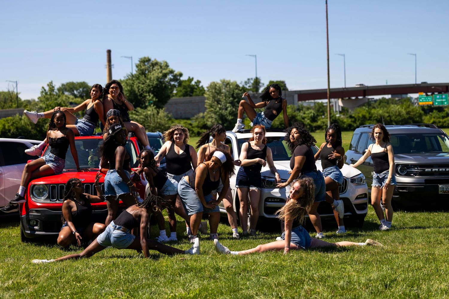 A large group of young women posing together on cars in a sunny outdoor parking area on a bright summer day.