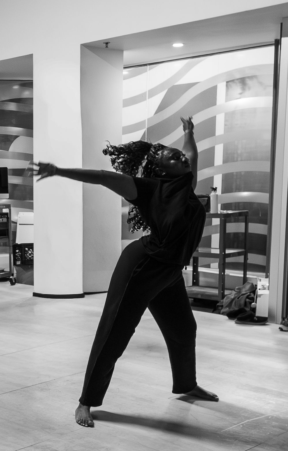 Black and white photo of a woman dancing gracefully in a black outfit, arms extended, in a modern indoor space.