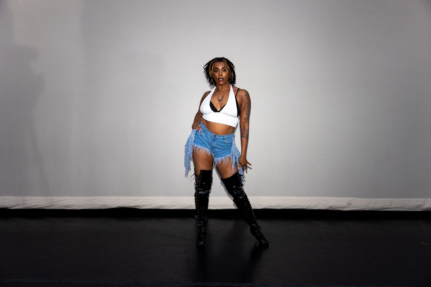 A dancer poses in white crop top, denim fringe shorts, and black thigh-high boots against a gray studio wall.