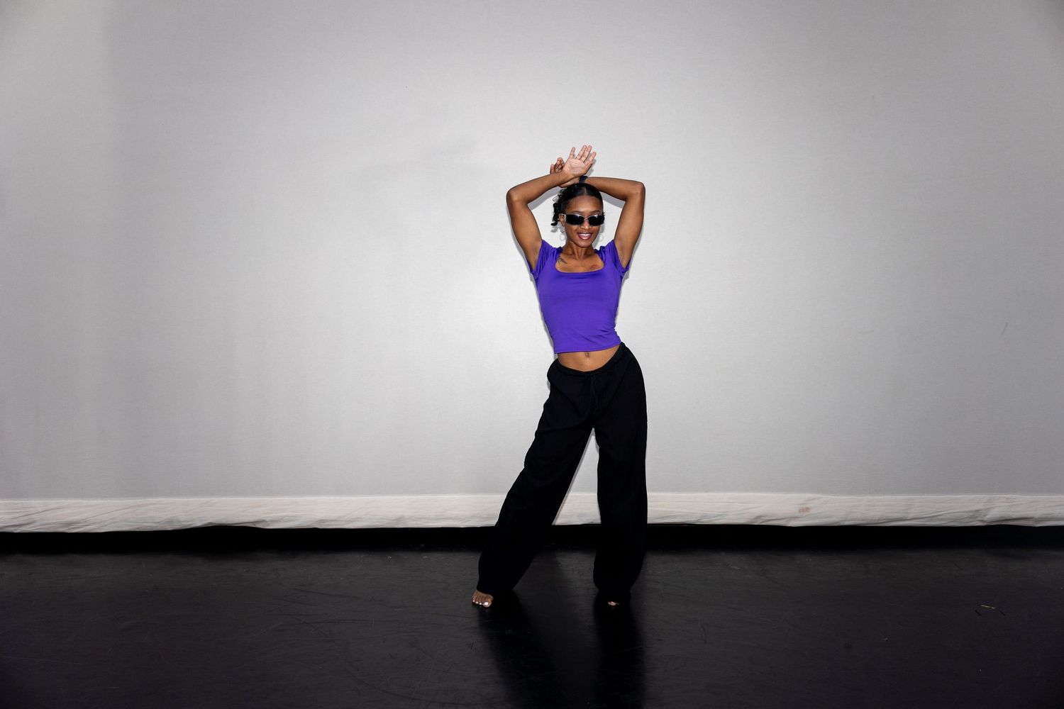 Black woman in purple crop top raises arms overhead while dancing in studio setting.