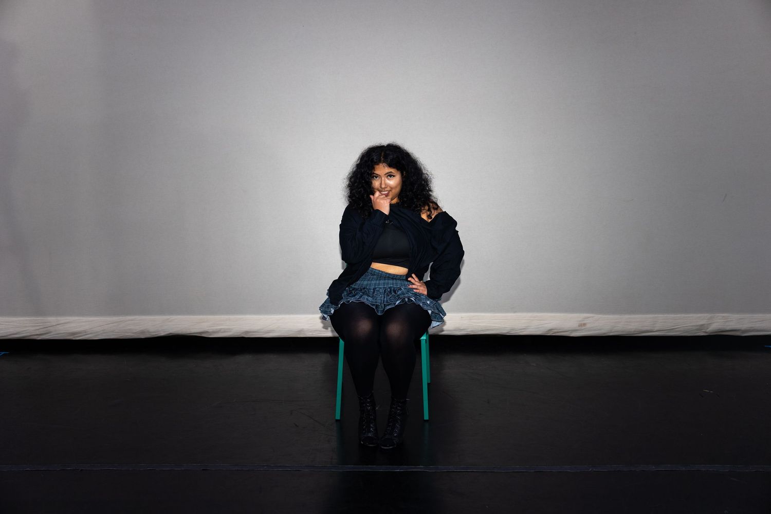 Young woman in denim skirt and dark jacket sits thoughtfully on green chair against gray studio wall.
