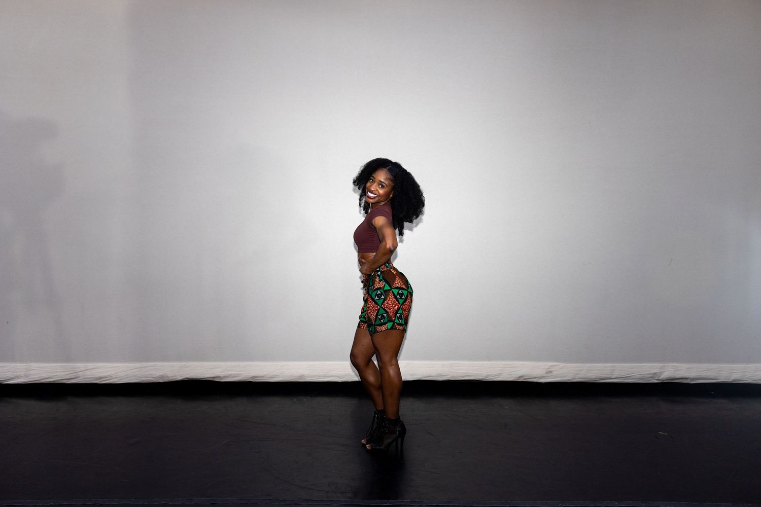 A woman in a colorful printed two-piece outfit poses sideways showing off her figure against a gray background.