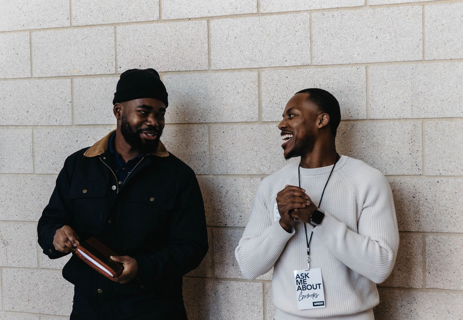 Two friends share a lighthearted moment while standing against a white brick wall.
