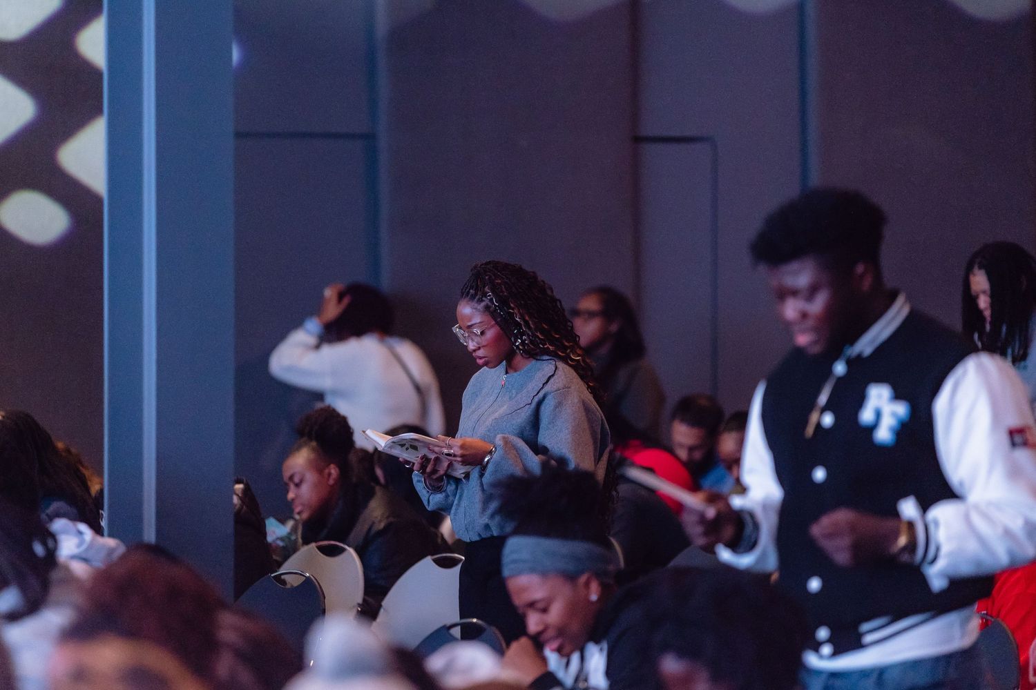 Audience members sit in rows watching a performance in a darkened theater or auditorium.
