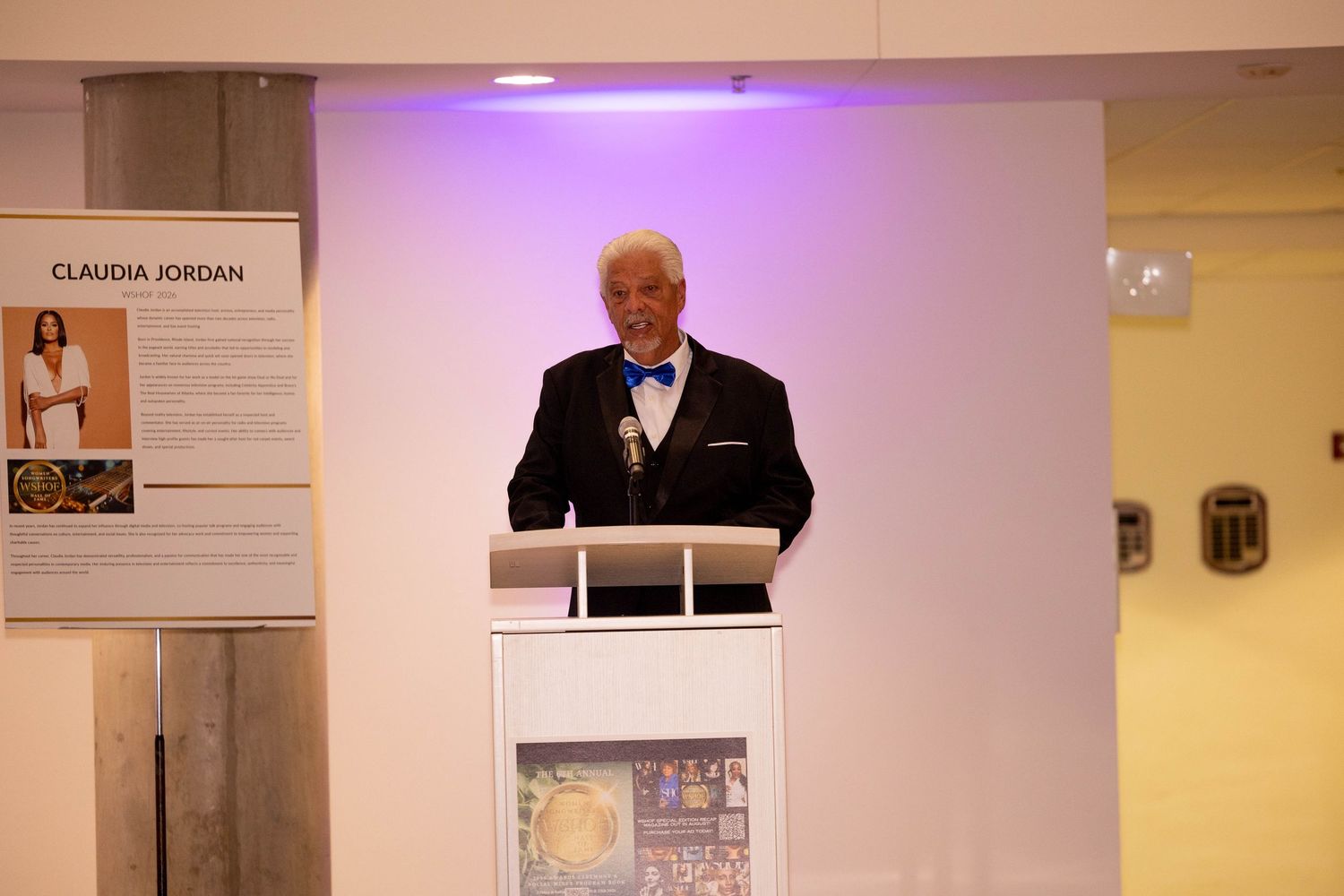 Elegant man in tuxedo with blue bow tie speaks at white podium during formal event near Claudia Jordan display.