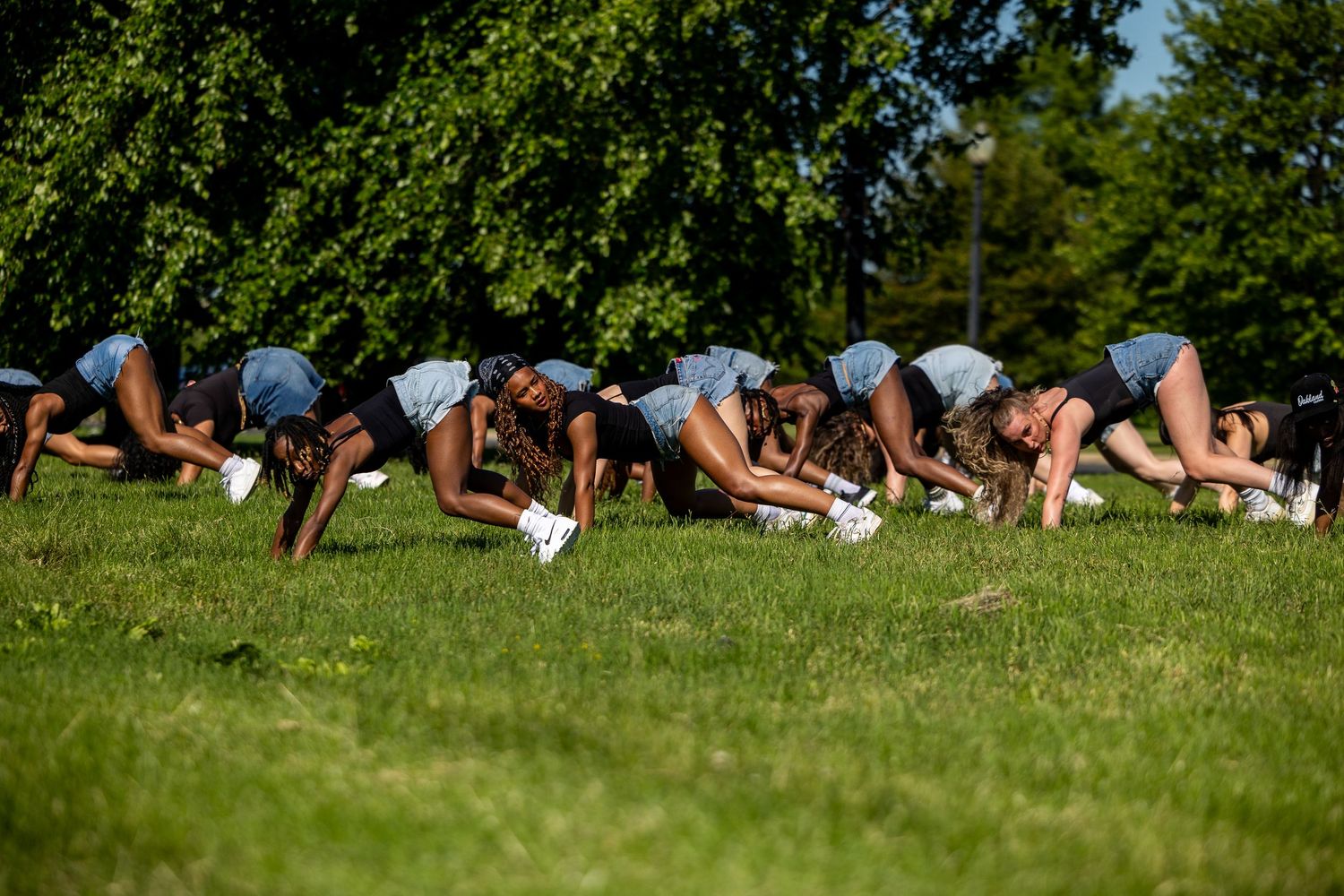 Group of young athletes doing push-ups on green grass in a sunny park during outdoor sports training session.