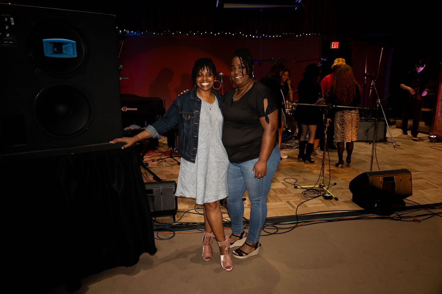 Two women pose together smiling near a speaker at an indoor live music venue.