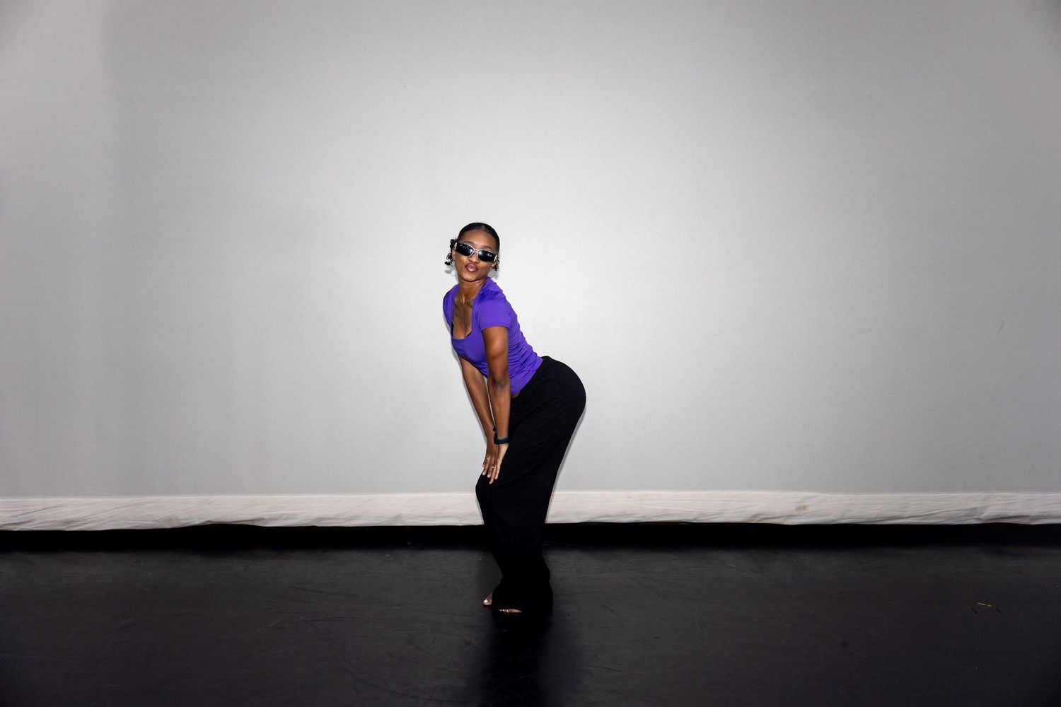 A woman in a purple top and black pants strikes a dynamic pose wearing sunglasses in a dance studio.