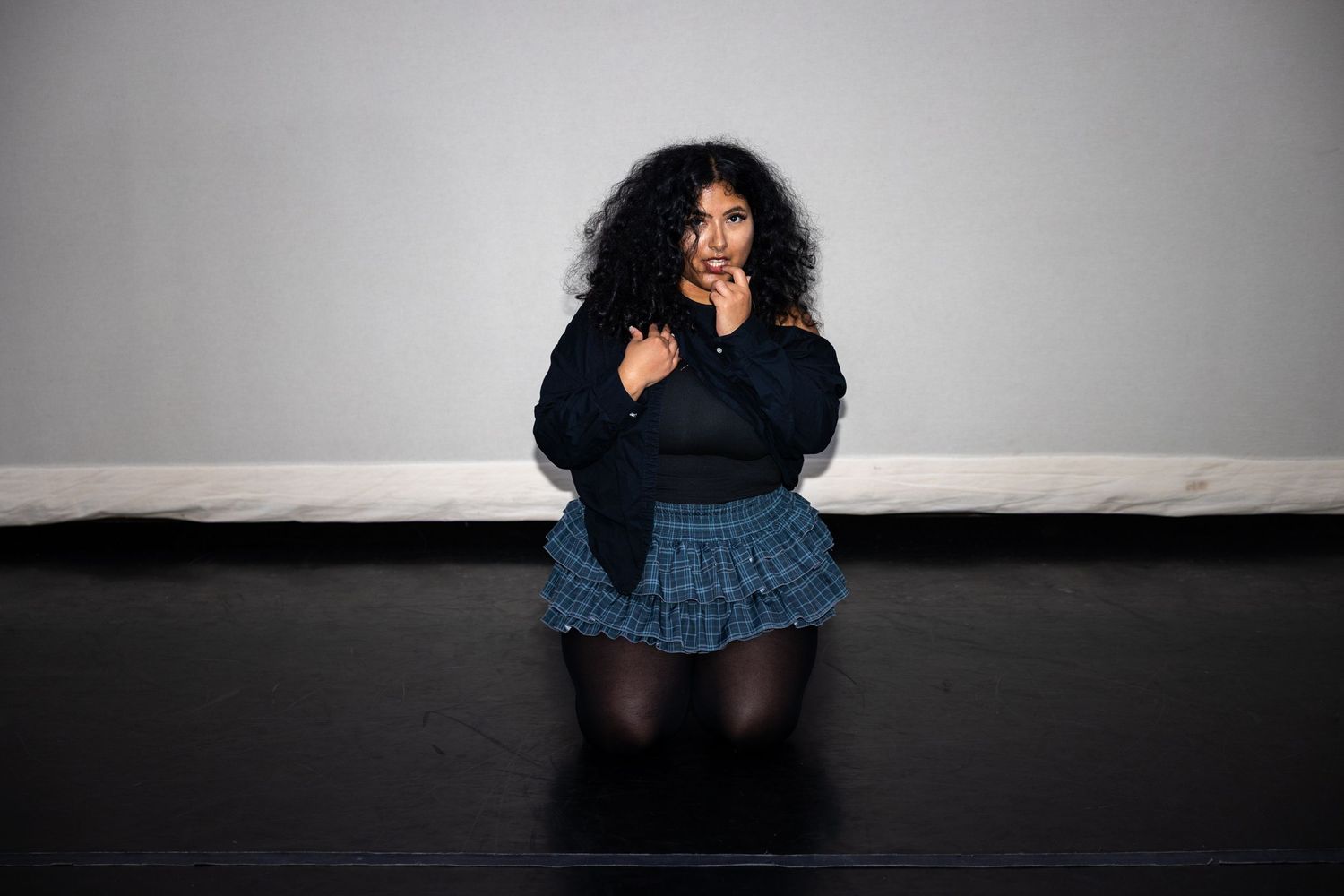 Curly-haired woman sitting on black studio floor in black sweater and blue plaid skirt, posing.
