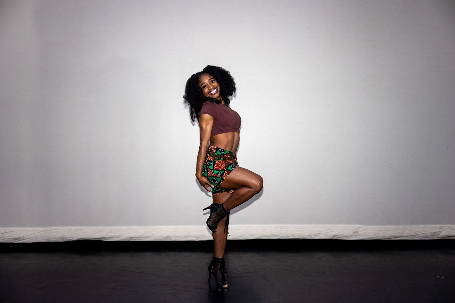 A smiling dancer in a colorful African print skirt and crop top poses on one leg.