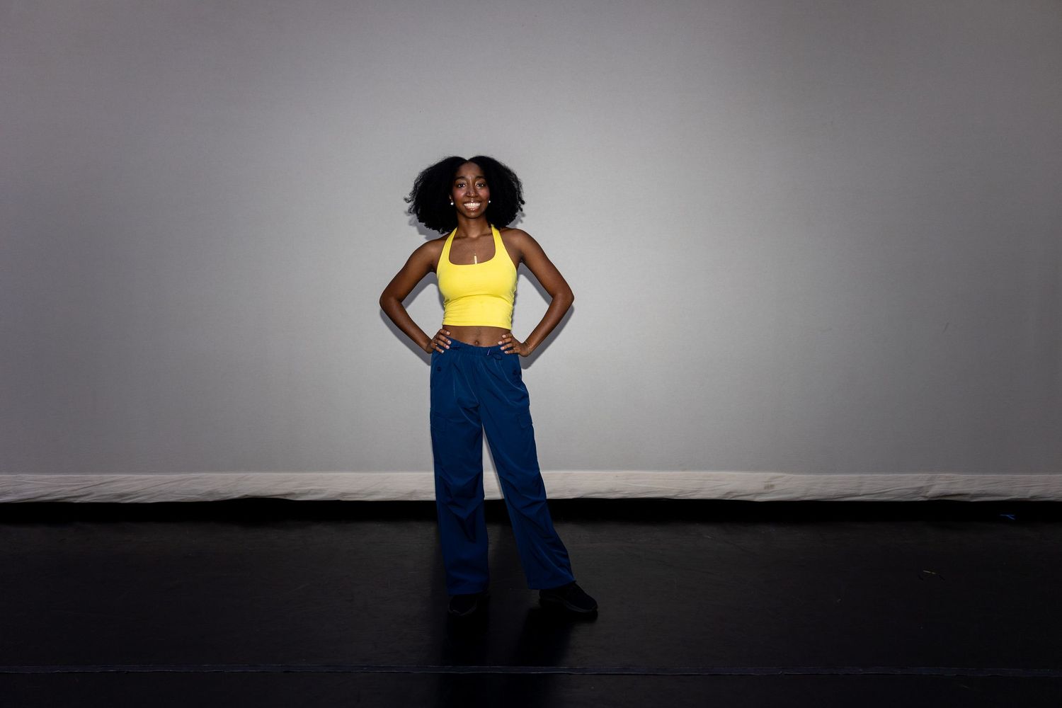 Confident woman in yellow crop top and blue wide-leg pants posing against gray studio background.