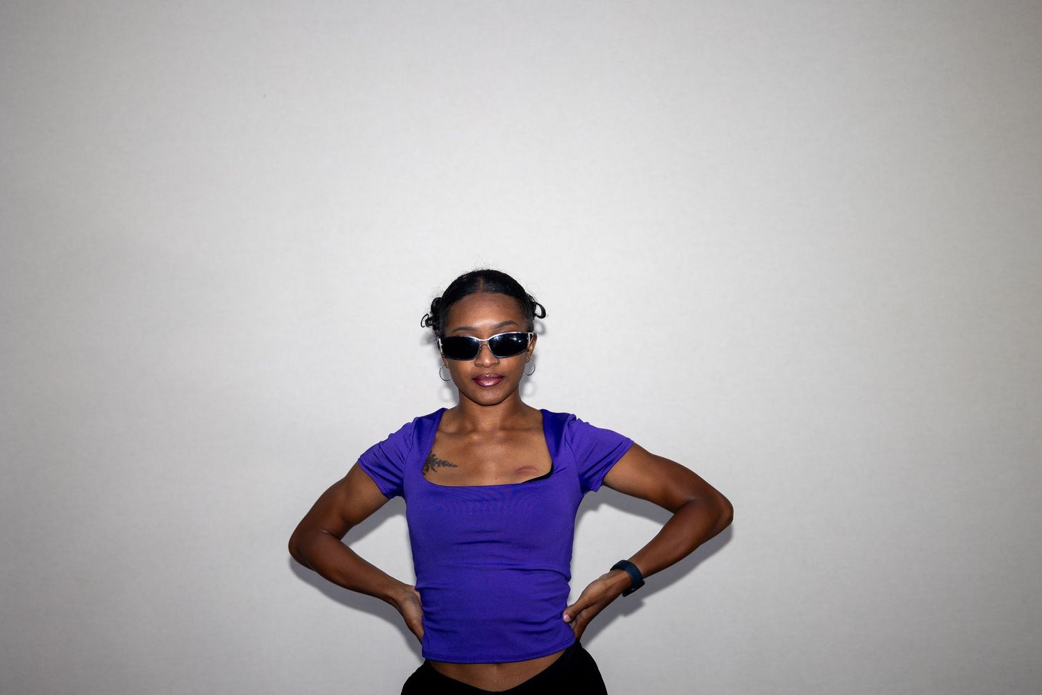 Confident young woman in purple crop top and sunglasses posing with hands on hips against white background.