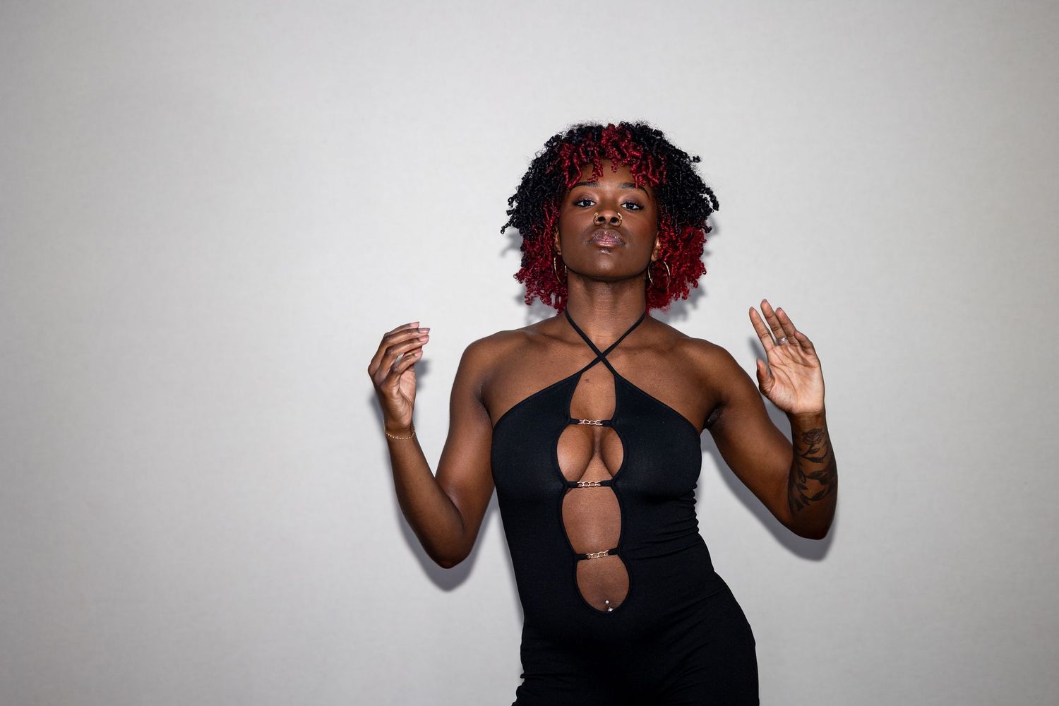 Woman with red curly hair wearing a black cutout dress, posing with eyes closed and hands raised gracefully.