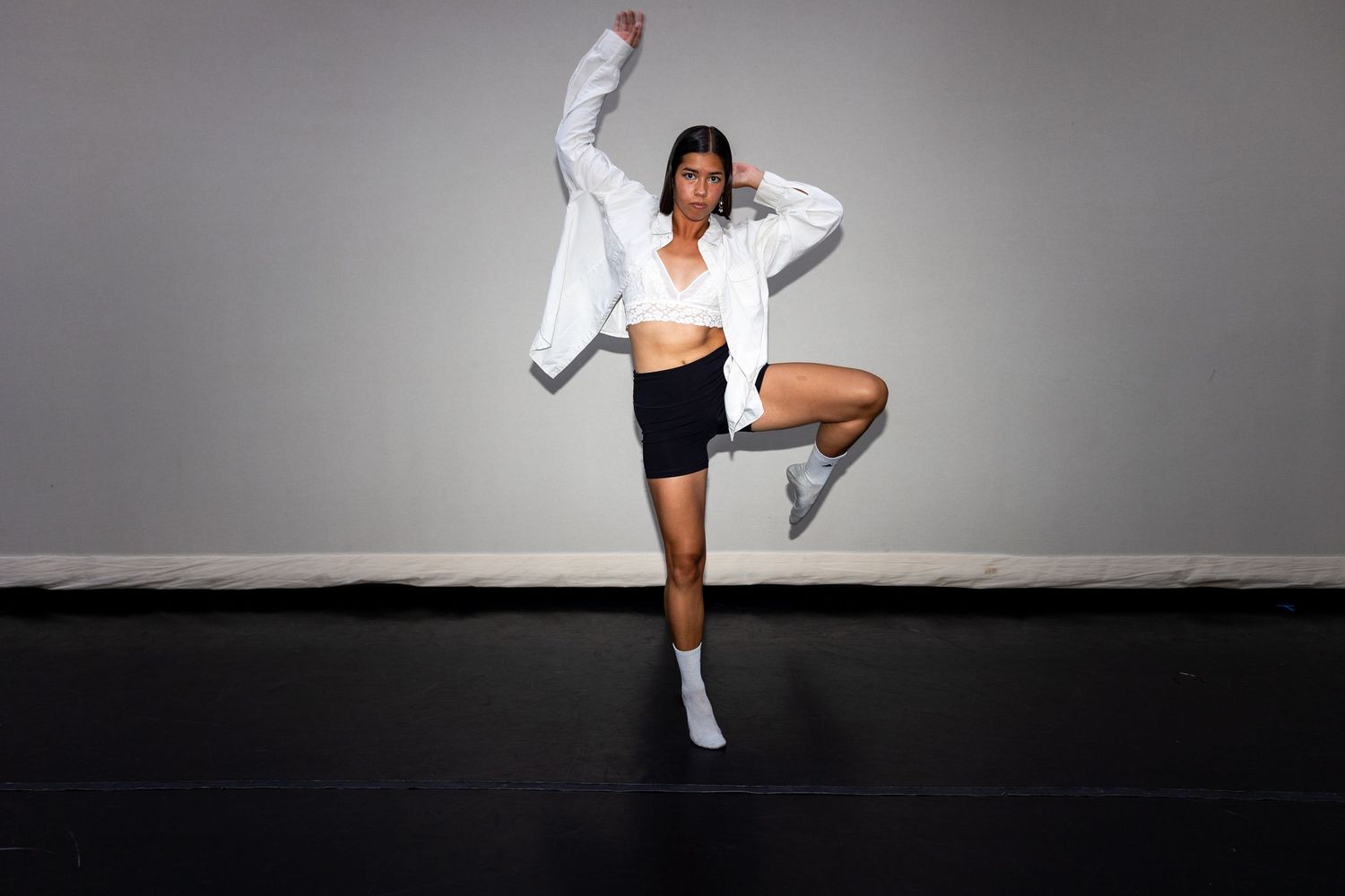 A dancer in a white open shirt and black shorts balances on one leg in a studio.