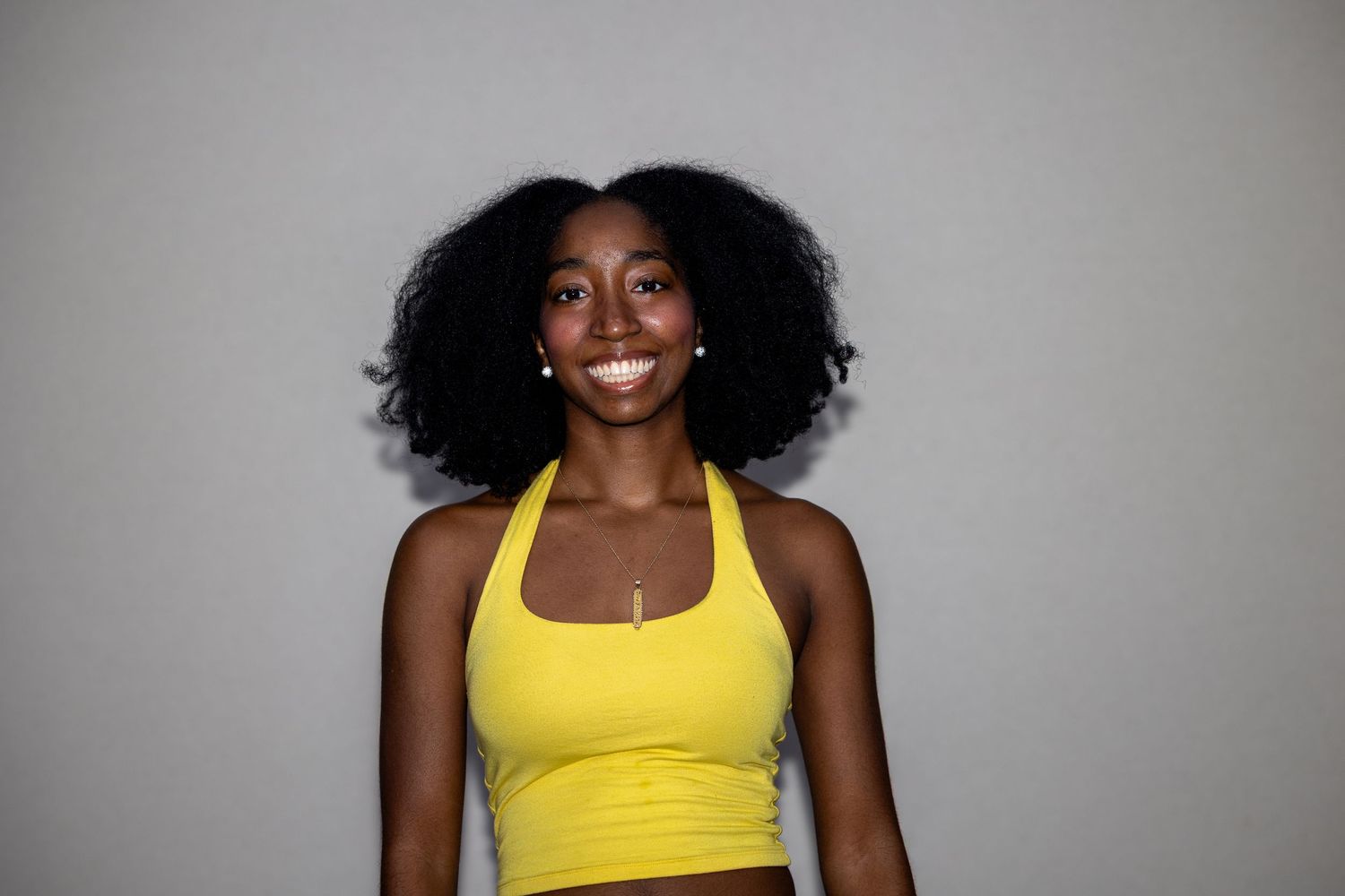 Smiling young Black woman with natural afro hair wearing a yellow crop top against a gray background.