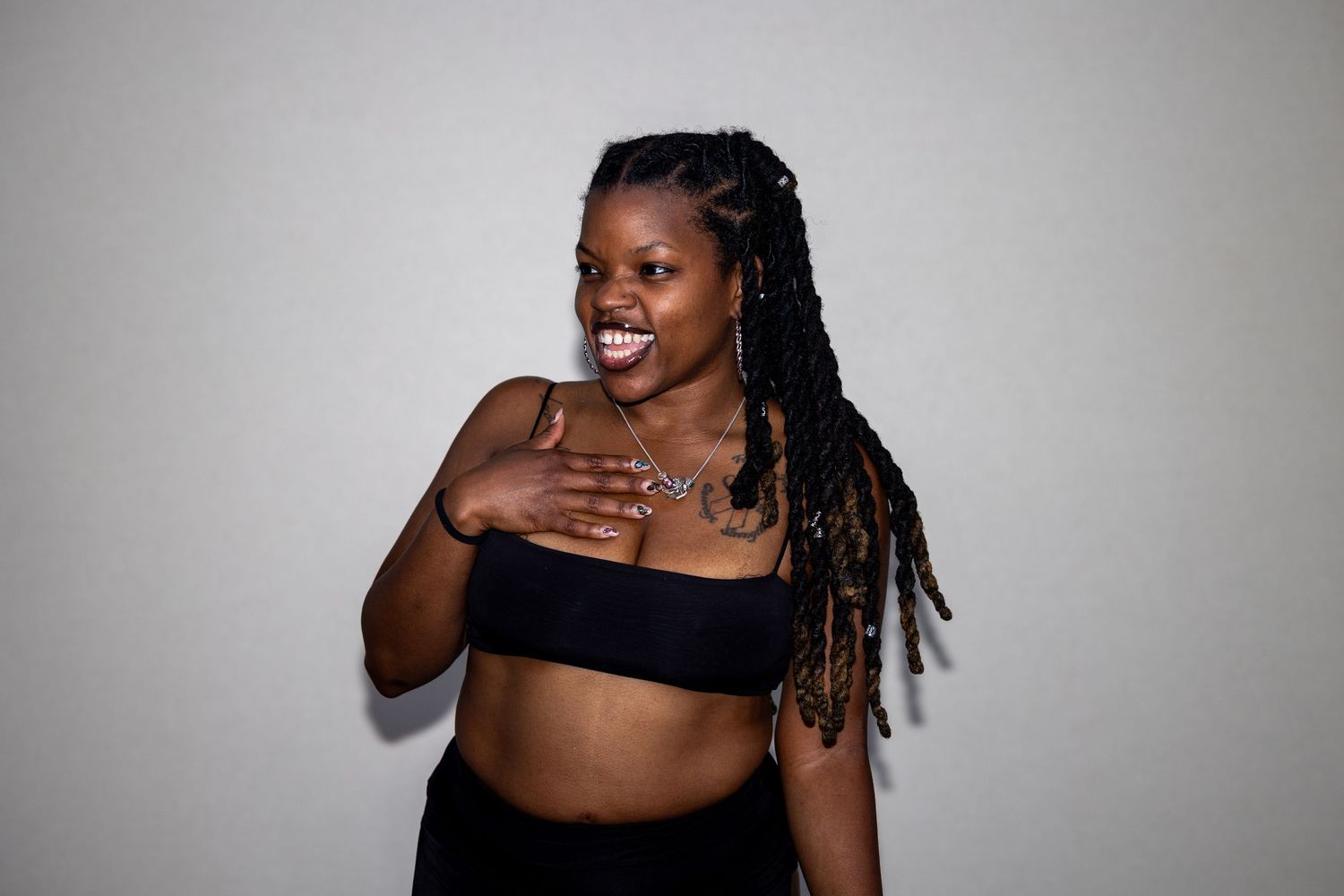 A smiling woman with braided locs wearing a black crop top poses with hand on chest against a gray background.
