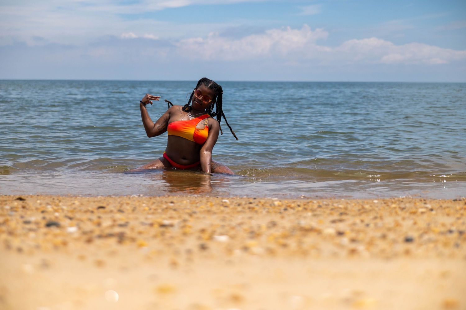 A woman with braids in a colorful bikini sits in shallow ocean waves on a sandy beach under a partly cloudy sky.