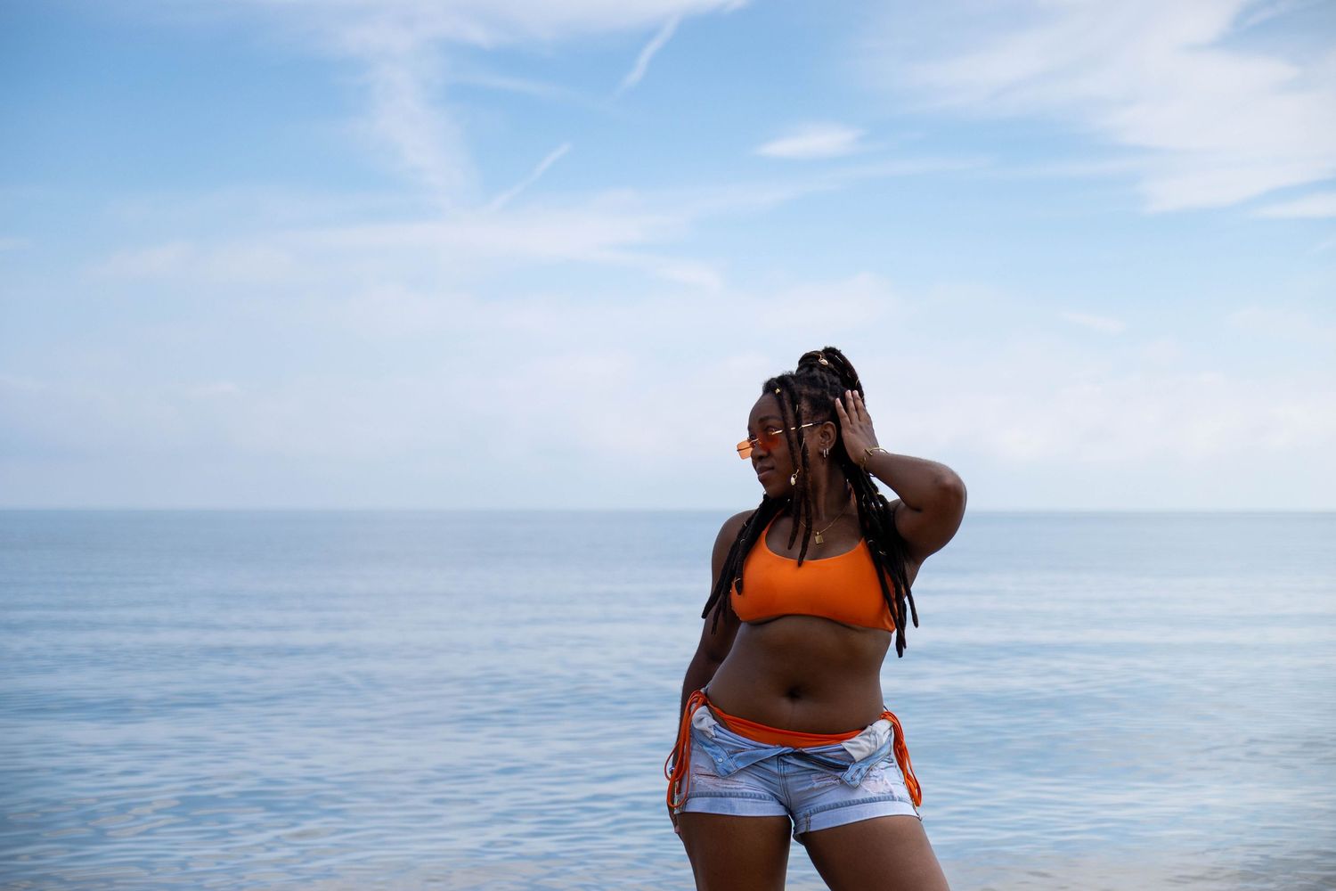 Young woman in orange bikini top and denim shorts standing by the ocean on a bright sunny day at the beach.