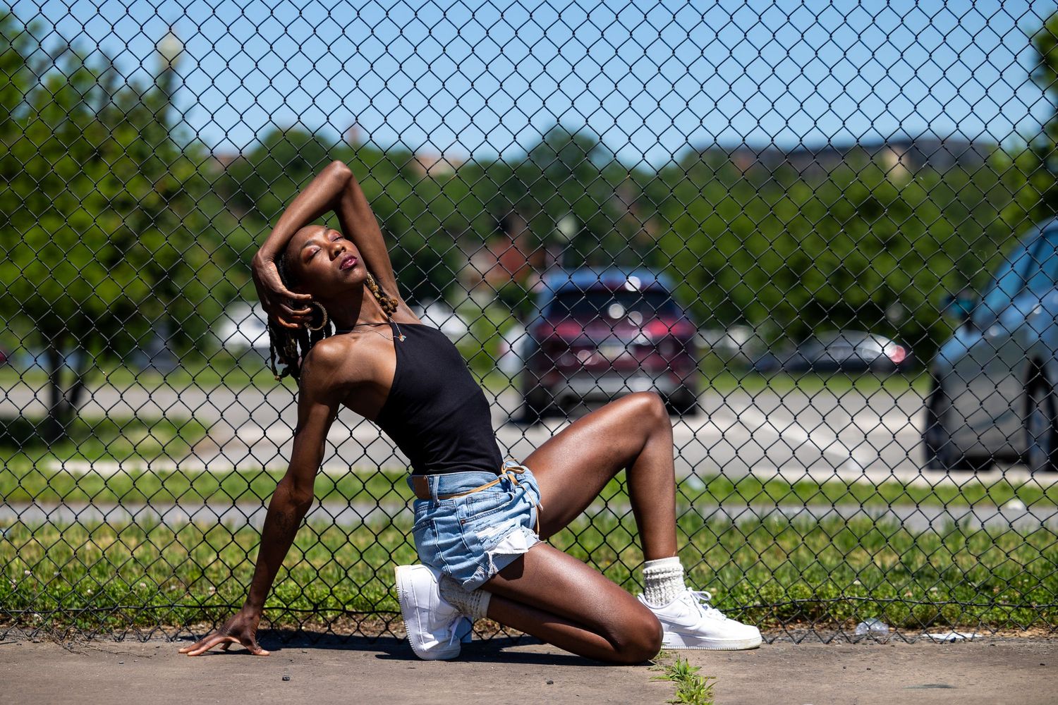 Young woman posing gracefully by a chain-link fence outdoors on a sunny day in denim shorts and sneakers.