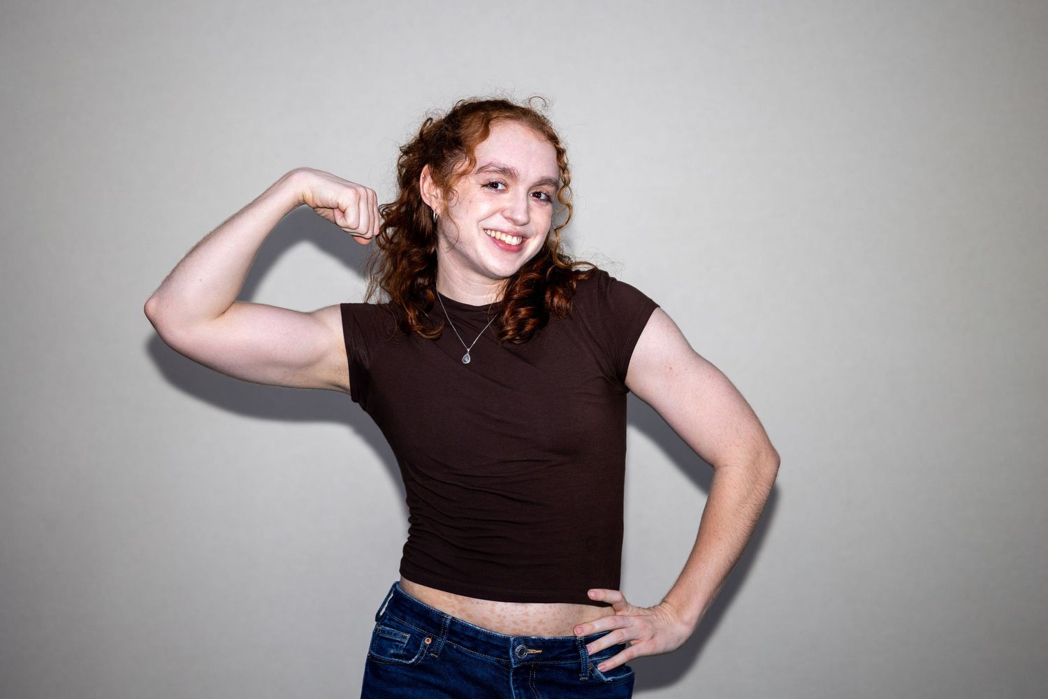 Young woman with curly red hair wearing a brown crop top flexing her arm and smiling against a gray background.